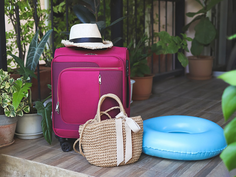 Front view of travel accessories, pink luggage, woven bag, backpack, headphones, swimming ring  and straw hat on balcony with houseplant.