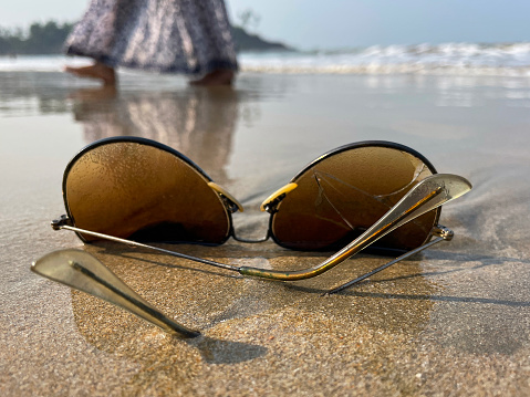 Stock photo showing close-up view of sandy beach at low tide with a pair of aviator, metal framed, tinted, mirrored sunglasses washed up from polluted sea.