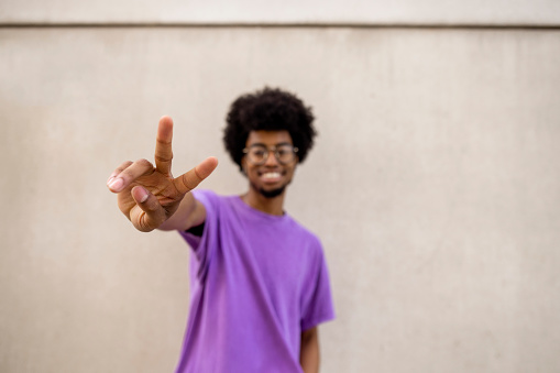 A front-view waist-up portrait shot of a young man smiling in front of a wall in the city wearing casual clothing on a summers day in Newcastle, England. He is looking at the camera and making a peace sign.