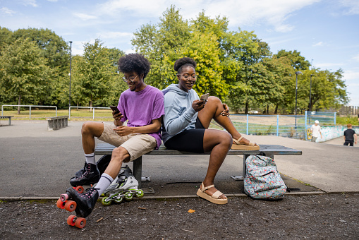 A shot of a young man and woman sitting down on a bench at a public park in the city, they are wearing casual clothing and shorts in the sun on a summers day in Newcastle, England. They are putting their skates on and relaxing while using their smartphones before they start skating.