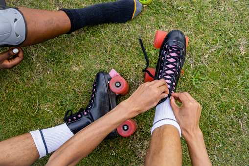 A point of view shot of an unrecognisable young man and woman sitting down on the grass at a public park in the city, they are putting rollerskates and knee pads on on a summers day in Newcastle, England.