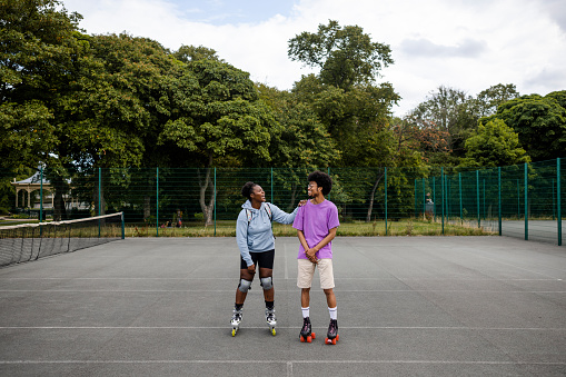 A front view shot of a young man and woman standing wearing rollerblades and rollerskates next to each other at a public park tennis court in the city, the young man is teaching his friend how to rollerblade on a summers day in Newcastle, England.