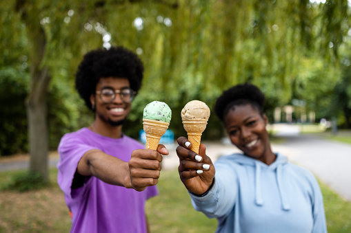 A front view shot of a young man and woman wearing casual clothing standing together showing ice creams to the camera in a public park on a summers day in Newcastle, England.