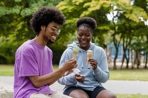 A front view shot of a young man and woman wearing casual clothing sitting together on a bench enjoying ice creams in a public park on a summers day in Newcastle, England.