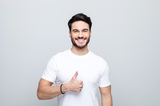Happy handsome man wearing white t-shirt standing with thumb up and laughing at camera. Studio shot, grey background.