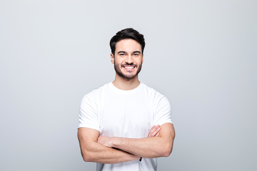 Happy handsome man wearing white t-shirt standing with arms crossed and smiling at camera. Studio shot, grey background.