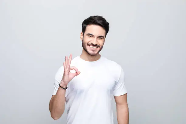 Happy handsome man wearing white t-shirt showing ok sigh and laughing at camera. Studio shot, grey background.