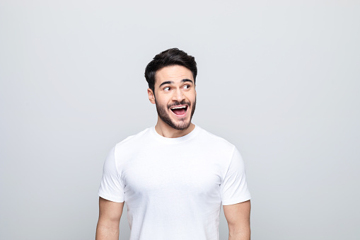 Excited handsome man wearing white t-shirt looking away and laughing. Studio shot, grey background.