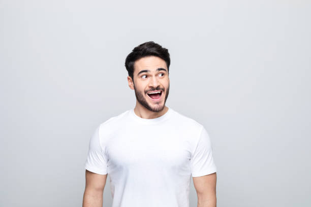 Portrait of surprised young man Excited handsome man wearing white t-shirt looking away and laughing. Studio shot, grey background. surprised men stock pictures, royalty-free photos & images