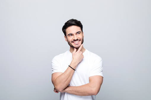 Handsome man wearing white t-shirt standing with hand on chin, looking away and smiling. Studio shot, grey background.