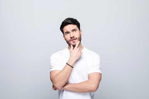 Handsome man wearing white t-shirt standing with hand on chin and looking away. Studio shot, grey background.