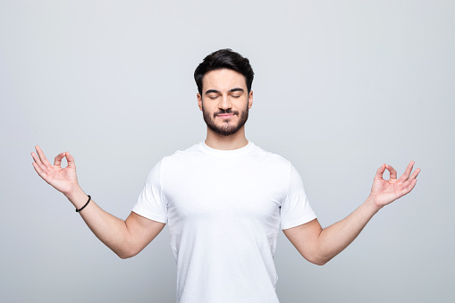 Happy handsome man wearing white t-shirt standing with raised hands in meditation pose. Studio shot, grey background.