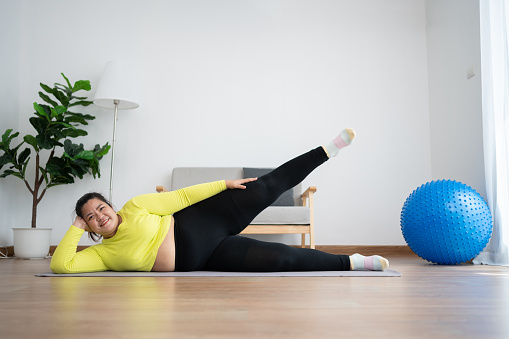 Asian overweight woman doing stretching exercise at home on fitness mat. Home activity training, online fitness class. Stretching training workout on yoga mat at home for good health and body shape.