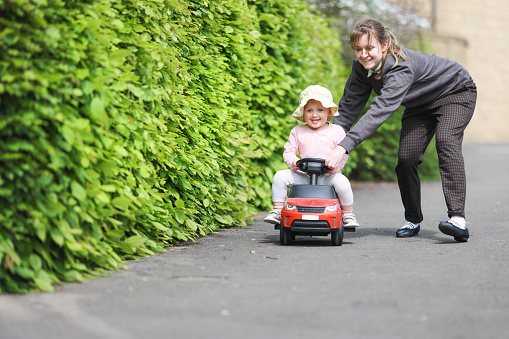 Young toddler laughing as she zooms down a residential path sitting on a toy car being pushed along by her mum.