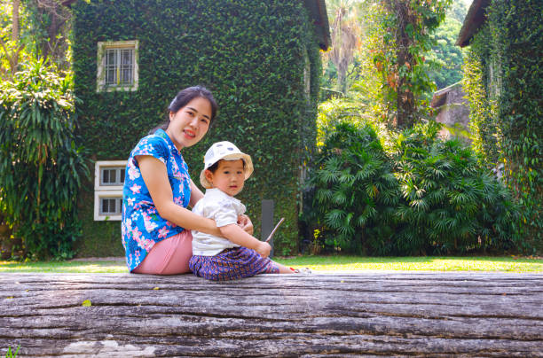 Portrait of Asian mother and son smiling and looking at camera while sitting on big log with greenery creeper plant on house wall in natural background