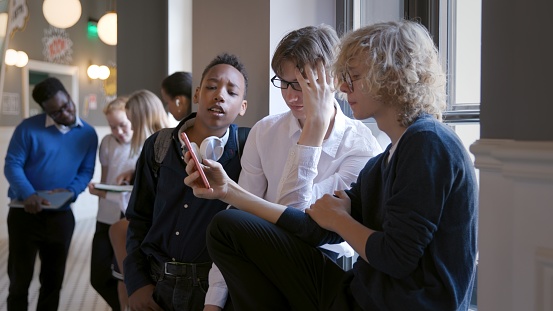 Multiracial young friends using mobile phone while sitting on windowsill in school corridor. Happy teenage students from different cultures sharing content on social networks