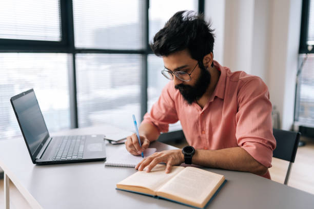 istock Concentrated bearded Indian businessman in glasses using laptop analyzing working project results, writing notes siting at table by window. 1490634873
