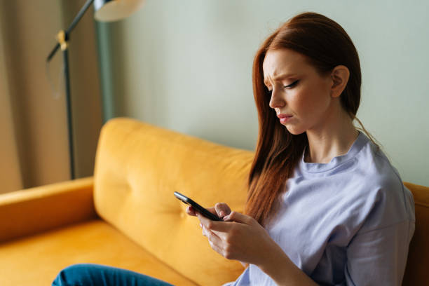 istock Sad red-haired young woman looking at smartphone screen expression face thinking about problem sitting on yellow couch, difficulty, feeling failure 1490634926
