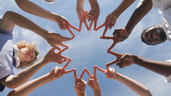 Bottom view of multiethnic teen students making finger circle outdoors. Happy diverse teenagers with teacher join hands together in circle over blue sky background