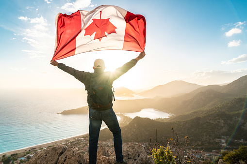 A Celebration of National Pride. Man holds waving national flag of Canada standing at the top at sunset. Beautiful mountains and sea on background.