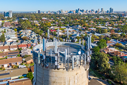 Aerial view, looking down on top of vertical concrete water tank providing telecommunications tower in an urban landscape: wide array of telecommunications equipment attached to its top providing communications services (including 5G, 4G) to urban residential and business districts in eastern suburbs of Adelaide, South Australia. The treelined streets are displaying remnant autumn colours. In the background is the Adelaide CBD skyline.
