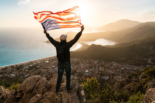 A Celebration of National Pride. Man holds waving national flag of USA standing at the top at sunset. Beautiful mountains and sea on background.