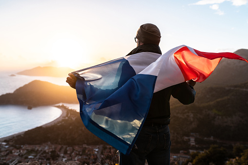 A Celebration of National Pride. Man in national flag of France standing at the top at sunset. Beautiful mountains and sea on background.