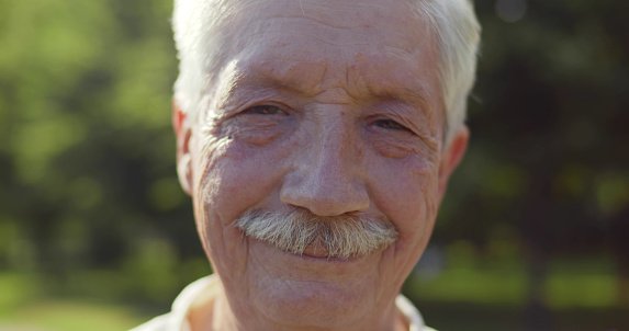 Bokeh close up portrait of happy senior man looking at camera outdoors. Cheerful elderly grandfather with mustache smiling at camera over blurred summer park background