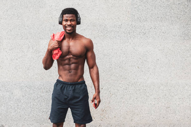 un homme afro-américain sportif se tient contre le mur sans t-shirt et sourit, un gars athlétique avec un corps attrayant écoute de la musique et sourit - male torso abdominal muscle the human body photos et images de collection