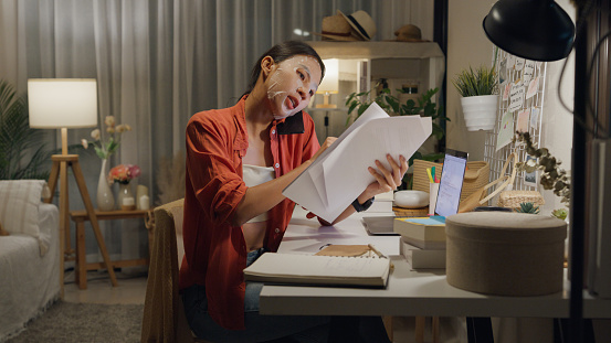 Young Asian woman use mask sheet sit front of desk with computer laptop and using mobile phone talk with colleague meeting work in living room at house at night. Work from home concept.