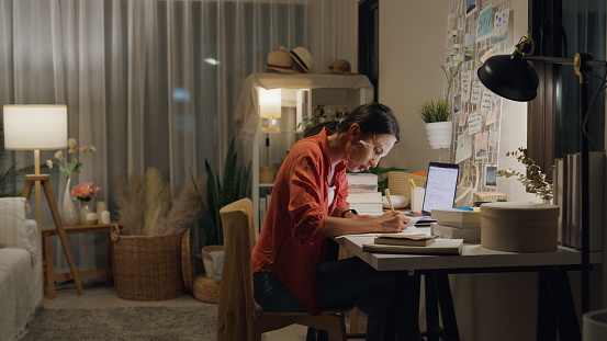 Young Asian woman use mask sheet on face sit front of desk with computer laptop and think create on work idea in living room at house at night. Work from home concept.