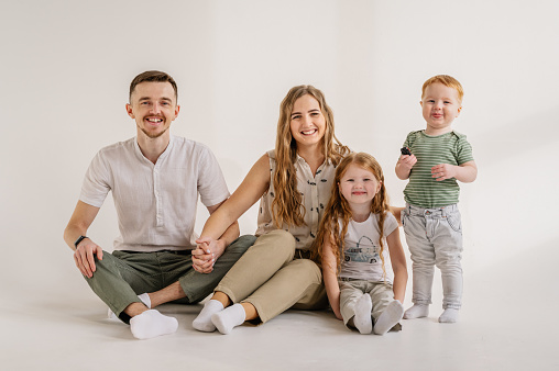 Young happy mother, father with child daughter, son isolated on white background. Funny parents hugs children sit, relaxing on floor near wall at home. Family moving into new house. Relocation concept