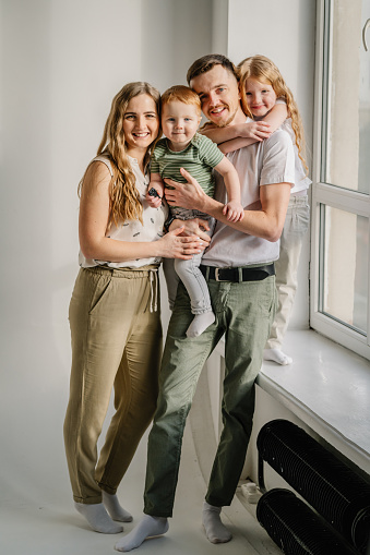 Young happy mom, dad with child daughter, son on white background studio near window. Mother and father playing active games with kids girl, boy. Funny parents hugs children relaxing on wall at home.