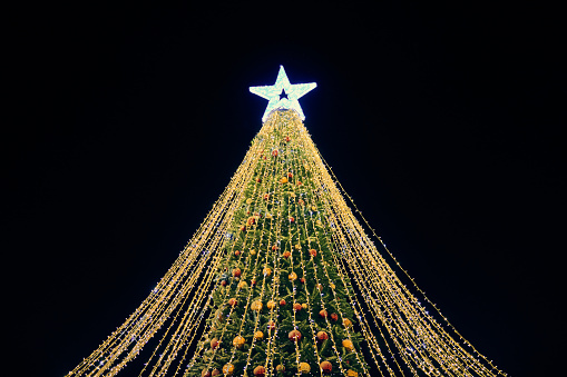 Christmas tree with yellow garlands, decorative bulbs and big white star topper at night blue sky background, outdoor holiday atmosphere. Festive Christmas tree with luminous yellow light garlands