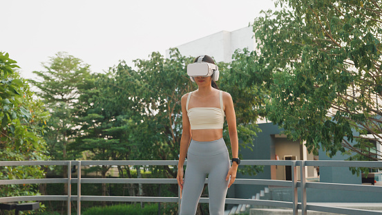 Young Asian athletic woman wearing virtual reality headset sportive top and leggings practicing yoga outdoors in city park. Diet and healthy weight loss concept.