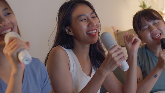 Young Asian female friends singing karaoke having fun at colorful house party at night. Lifestyle together.