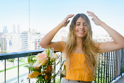 portrait of young latin woman of colombian ethnicity, dressed in yellow, sitting on her apartment balcony, confused with hands on head, smiling and looking at camera, lifestyle concept, copy space.