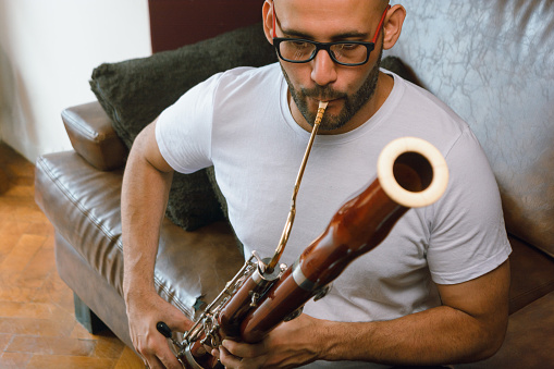 front view of latino man of venezuelan ethnicity, with beard, bald head and glasses, sitting on the sofa in the living room of the house concentrated playing the bassoon reading sheet music.