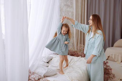 Charming mother and daughter in casual clothes of pale blue color have fun in the bedroom on the bed. Depict dance. Happy childhood concept.