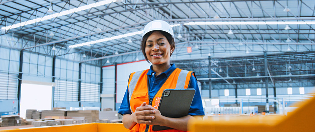 Portrait of black female engineer technician smiling, wear hardhat, holding digital tablet in warehouse packaging factory. Logistic industry business, industrial job, or people at work concept