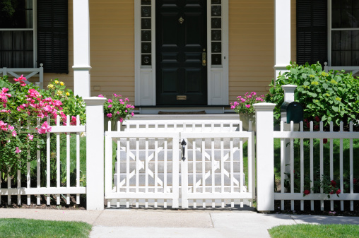 Little white gate and picket fence with roses. Frontal view, high contrast, porch and house facade in background
