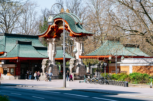A sunny day in Berlin, Germany. The main entrance to the Berlin Zoo is in the foreground, and people are strolling and passing by. The entrance is a beautiful archway with two elephants carved into the stone. The zoo is located in the Mitte district of Berlin, and it is one of the most popular tourist destinations in the city.