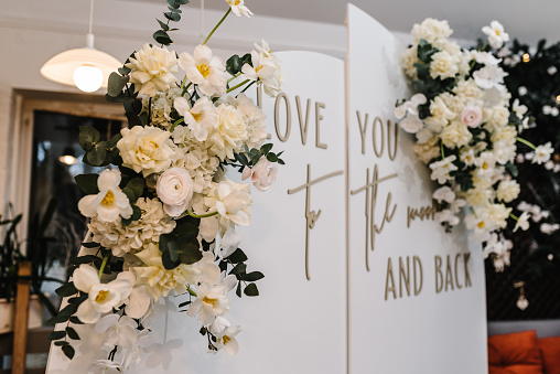 Arch decorated with flowers, and greenery with the text love you to the moon and back. Wedding reception for the luxury ceremony. Trendy decor party in the banquet area. Photo-wall white.