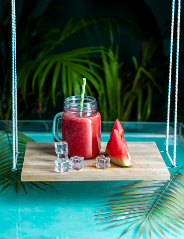 Mason jar with healthy watermelon juice and ice cubes, beautiful juice image hanging swing with green leaves and water