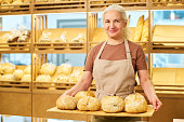 Happy mature female baker or clerk in apron holding wooden tray with bread