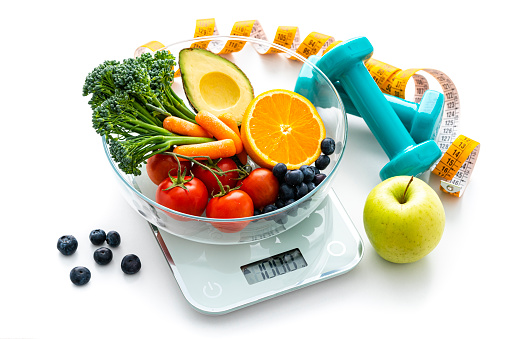 Healthy eating and exercising concept. High angle view of a glass bowl filled with fresh organic fruits and vegetables placed on a kitchen scale isolated on white background. Yellow tape measure and dumbbells complete the composition. The composition includes orange, carrot, broccoil green apple, tomatoes and berry fruits.