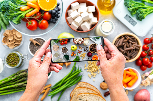 Woman's hands holding smartphone photographing a table filled wit a large group of healthy fruits, vegetables and soy products.