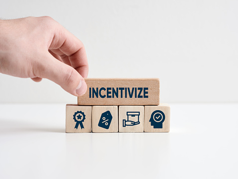 Incentivize concept. To motivate or encourage someone with an incentive to do something. Hand places wooden block with the word incentivize with business symbols.