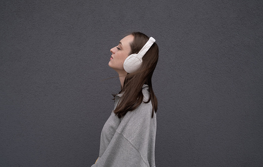 Carefree attractive brunette girl, dressed in a gray sweater, listening to music on headphones, standing on a gray background