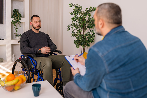 A young Caucasian man in a wheelchair engages in a deep conversation with a mature Caucasian psychotherapist. They sit in a comfortable and serene therapy room, creating a safe space for open dialogue